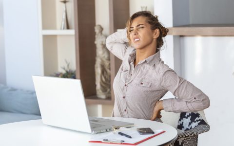 Portrait of young stressed woman sitting at home office desk in front of laptop, touching aching back with pained expression, suffering from backache after working on laptop